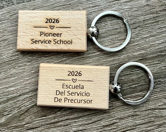 May include: Two rectangular light-colored wooden keychains with silver metal rings. The top keychain is engraved with 2026, a heart, and Pioneer Service School. The bottom keychain is engraved with 2026, a heart, and Escuela Del Servicio De Precursor.