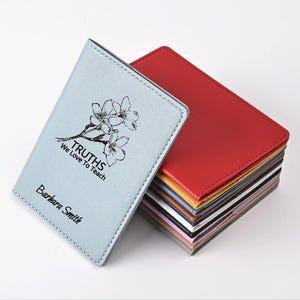 May include: A stack of colorful, leather-like wallets. The top wallet is light blue with the text "TRUTHS We Love To Teach" and "Barbara Smith" printed on it, along with a floral design. A red wallet is visible on top of the stack.