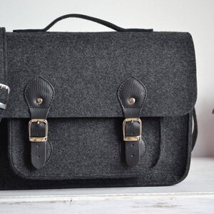May include: A dark gray felt messenger bag with two black leather buckles and a long strap. The bag has a flap closure and a small pocket on the front.