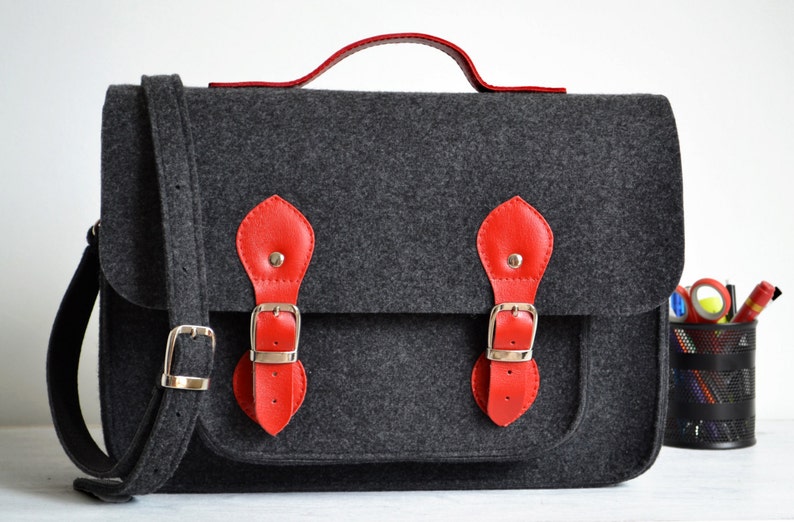 May include: A gray felt satchel bag with red leather accents and silver buckles. The bag has a top handle and a long adjustable shoulder strap.