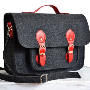 May include: A gray felt satchel bag with red leather accents and a black adjustable shoulder strap. The bag has two large buckles on the front and a smaller buckle on the strap.
