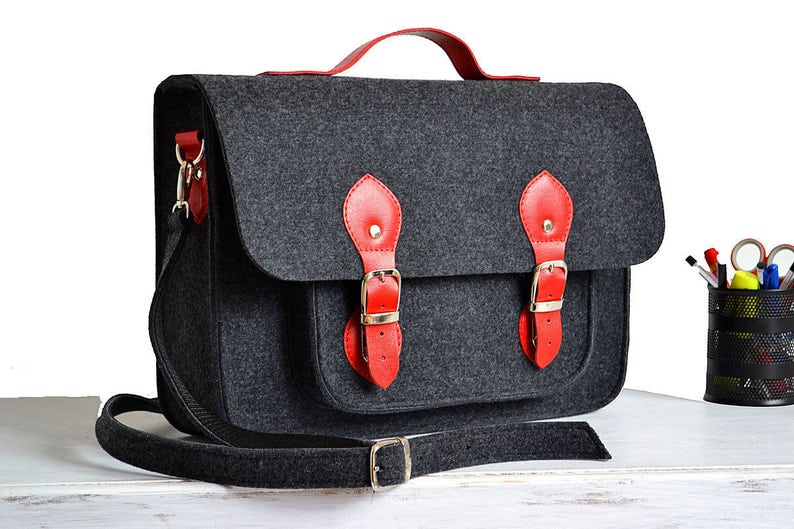 May include: A gray felt satchel bag with red leather accents and a black adjustable shoulder strap. The bag has two large buckles on the front and a smaller buckle on the strap.