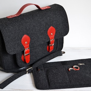 Puede incluir: Un bolso de mano de fieltro gris con detalles de cuero rojo y una bolsa de fieltro a juego. El bolso tiene un asa superior y una larga correa de hombro ajustable. La bolsa tiene varios bolsillos y una cremallera.