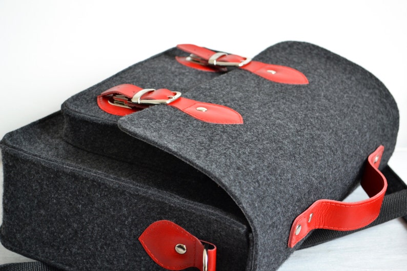 May include: A gray felt messenger bag with red leather accents and silver buckles. The bag has a top flap with a buckle closure and a red leather handle.