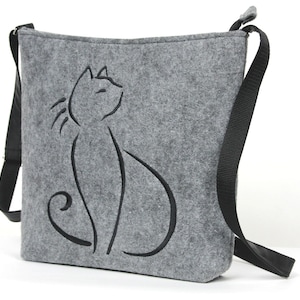 Puede incluir: Un bolso de hombro de fieltro gris con una correa ajustable negra. El bolso presenta un diseño bordado negro de un gato.