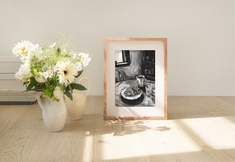 May include: A framed black and white sketch of a breakfast scene. A croissant and coffee cup sit on a table, next to a glass of water. A vase of white flowers and a pitcher are in the foreground.