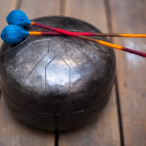 May include: A black steel handpan with two wooden mallets with blue and orange yarn wrapped around the ends. The mallets are laying on top of the handpan.