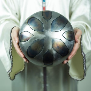 May include: A person holding a silver handpan with a floral design. The handpan has a dark gray finish and is slightly dented.