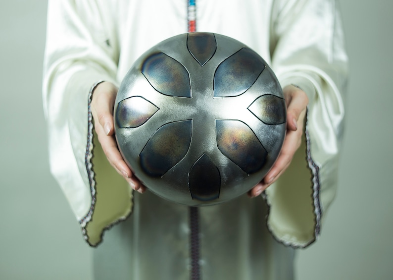 May include: A person holding a silver handpan with a floral design. The handpan has a dark gray finish and is slightly dented.