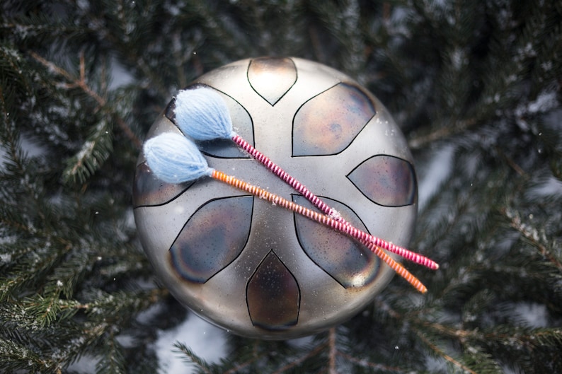 May include: A silver-colored handpan with a decorative pattern, resting on a bed of pine needles and snow. Two red and orange striped mallets with blue felt heads are resting on the handpan.