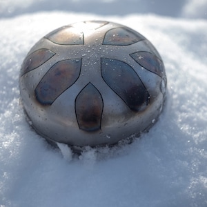 May include: A silver-colored handpan with a starburst design sits on a bed of white snow. The handpan has a slightly textured surface and appears to be made of metal.