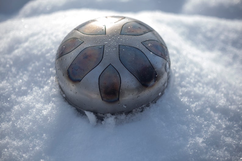 May include: A silver-colored handpan with a starburst design sits on a bed of white snow. The handpan has a slightly textured surface and appears to be made of metal.