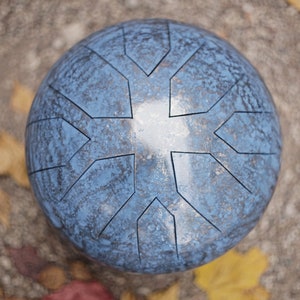 Op de afbeelding: Een blauwe stalen handpan met een geometrisch ontwerp. De handpan ligt op een bed van gevallen bladeren.