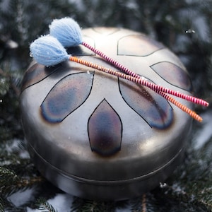 May include: A silver steel handpan with a snowflake design and two red and orange mallets with blue felt tips.