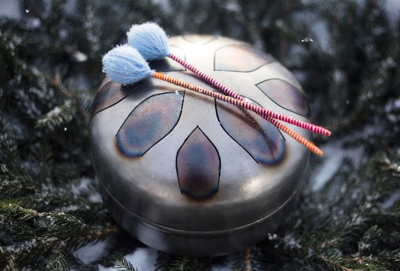 May include: A silver steel handpan with a snowflake design and two red and orange mallets with blue felt tips.