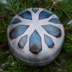 May include: A silver-colored handpan with a blue and brown design. The handpan is made of metal and has a circular shape. It is surrounded by green pine branches.