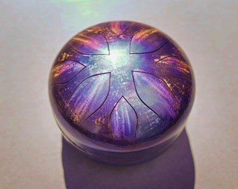 May include: A purple and gold handpan with a flower-like design. The handpan is a musical instrument that is played by tapping on the surface.