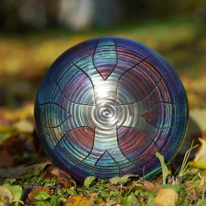 Könnte beinhalten: Eine Stahltrommel mit metallischem Finish in Blau-, Lila- und Rottönen. Das Instrument hat eine kreisförmige Form mit ausgeschnittenen Abschnitten und liegt auf einem Bett aus Blättern und Gras.