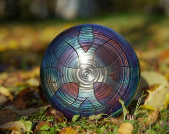 May include: A steel tongue drum with a metallic finish in shades of blue, purple, and red. The instrument has a circular shape with cut-out sections and is resting on a bed of leaves and grass.