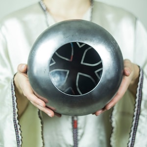 May include: A silver metal sphere with a circular opening revealing a black and white geometric design. The sphere is being held by a person's hands.