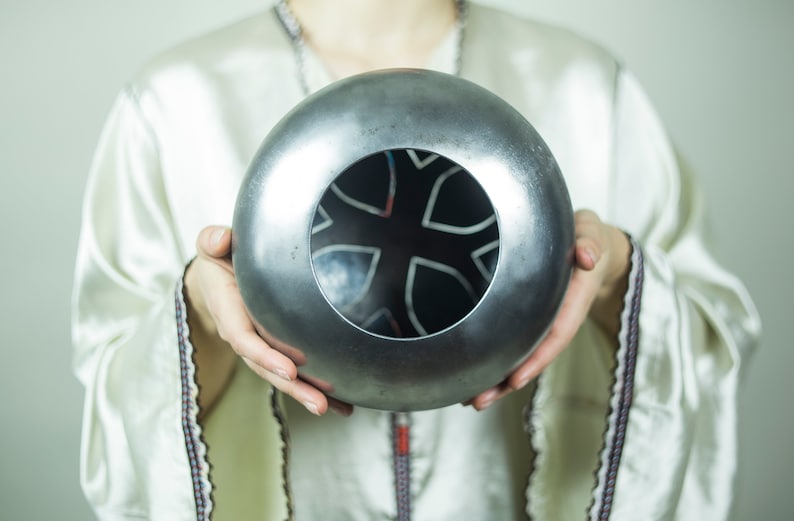 May include: A silver metal sphere with a circular opening revealing a black and white geometric design. The sphere is being held by a person's hands.