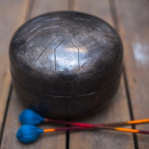 May include: A black steel handpan with a unique design, resting on a wooden surface. Two wooden mallets with blue and orange felt tips are lying next to the handpan.