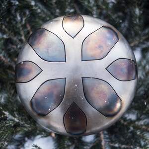 May include: A silver-colored handpan with a floral design. The handpan is surrounded by evergreen branches.