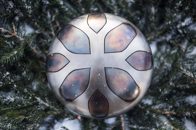 May include: A silver-colored handpan with a floral design. The handpan is surrounded by evergreen branches.