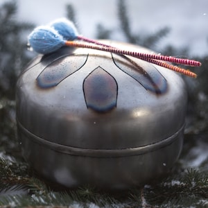 May include: A silver handpan with a unique design, resting on a bed of pine needles and snow. Two colorful mallets are resting on top of the handpan.