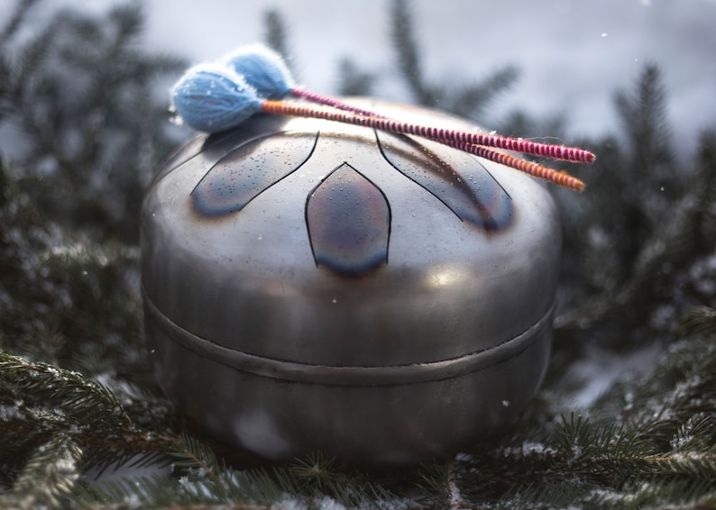 May include: A silver handpan with a unique design, resting on a bed of pine needles and snow. Two colorful mallets are resting on top of the handpan.
