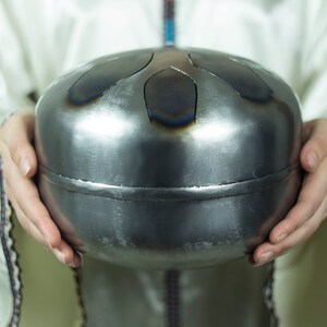 May include: A person holding a silver handpan with a circular shape and multiple indentations. The handpan is made of metal and has a smooth, shiny surface.