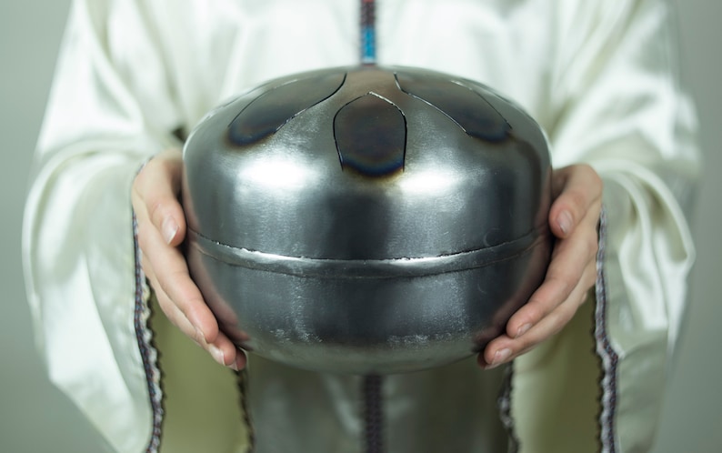 May include: A person holding a silver handpan with a circular shape and multiple indentations. The handpan is made of metal and has a smooth, shiny surface.