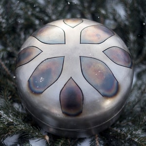 May include: A silver-colored handpan with a snowflake-like design. The handpan is surrounded by green pine branches and snow.