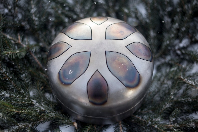 May include: A silver-colored handpan with a snowflake-like design. The handpan is surrounded by green pine branches and snow.