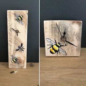 May include: A wooden clock with a rustic finish features three painted bumblebees. The clock has black hands and a gold center. The clock is standing on a wooden surface.