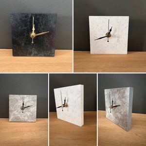 Puede incluir: Cuatro estilos diferentes de relojes cuadrados con caras de mármol negro, blanco y gris. Los relojes tienen manecillas negras y detalles dorados.