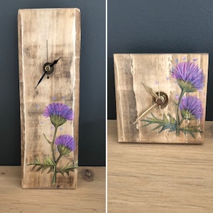 Peut inclure: Deux horloges en bois avec des fleurs de chardon pourpre peintes. Les horloges ont des aiguilles noires et des accents dorés.