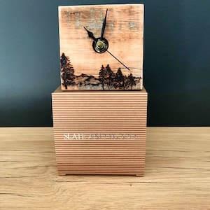 May include: A small wooden clock with a burnt wood design of a forest scene. The clock is on a brown box with the text "Slate and Wood" printed on the front.