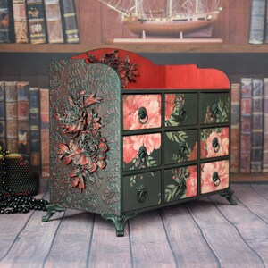 May include: A black wooden drawer organizer with a red top and floral patterned drawers. The organizer has decorative floral carvings and sits on four legs.