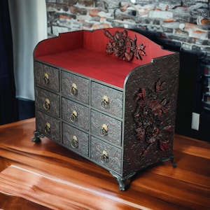 May include: A black wooden jewelry box with intricate floral carvings and a red velvet interior. The box has multiple drawers for storing jewelry or other small items.