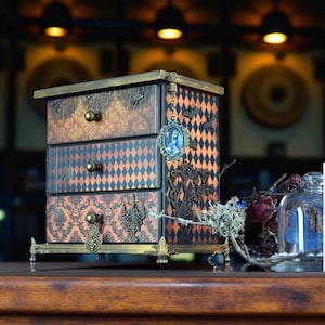 May include: A decorative jewelry box with three drawers, featuring a vintage design with orange and black diamond patterns. The box has ornate metal hardware and a hanging cameo pendant. Bats and a tree silhouette add to the gothic theme.