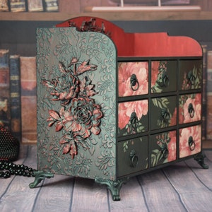 May include: A black wooden organizer with nine drawers featuring floral patterns in shades of pink and green. The organizer has decorative floral accents and sits on four ornate legs.