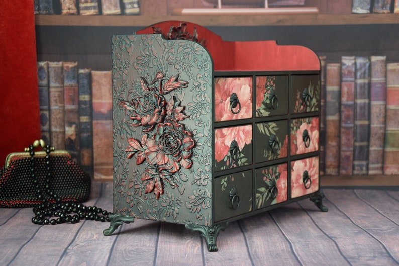 May include: A black wooden organizer with nine drawers featuring floral patterns in shades of pink and green. The organizer has decorative floral accents and sits on four ornate legs.