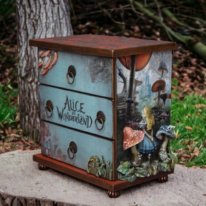 Pode incluir: Uma cômoda de três gavetas com o tema "Alice no País das Maravilhas". A cômoda apresenta um fundo azul claro com ilustrações da história. A parte superior é marrom avermelhado. O texto "Alice in Wonderland" é impresso nas gavetas.
