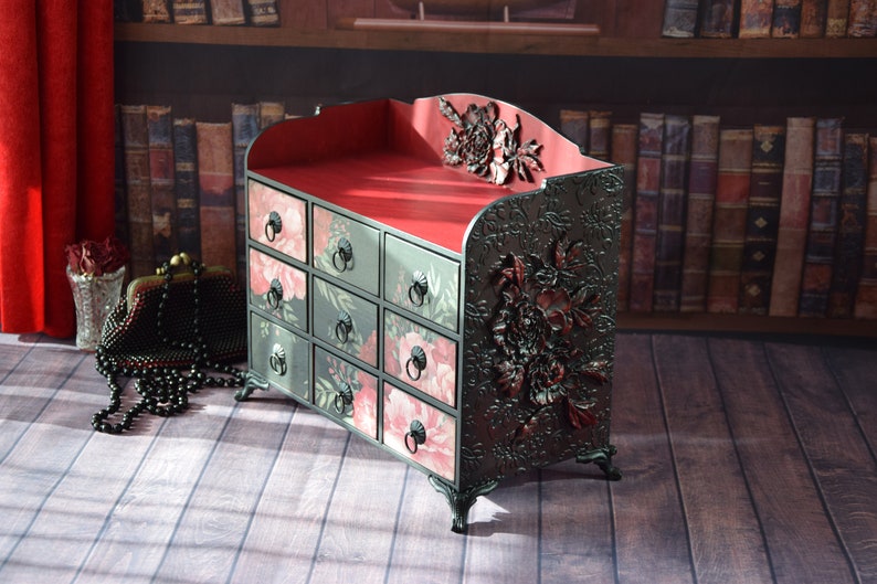 May include: A black wooden chest with 12 drawers, each with a floral design. The chest has a red velvet lining and ornate floral carvings on the sides.