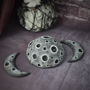 May include: Three grey, crescent moon-shaped sculptures with a textured surface and craters. The larger sculpture is a full moon with a hollow centre.
