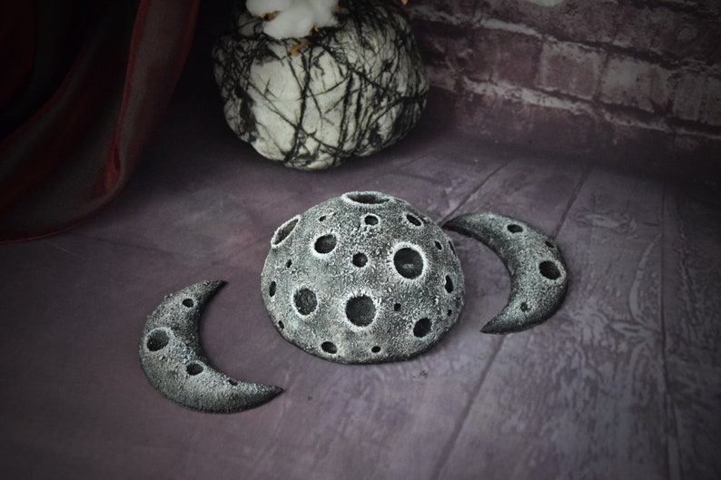 May include: Three grey, crescent moon-shaped sculptures with a textured surface and craters. The larger sculpture is a full moon with a hollow centre.