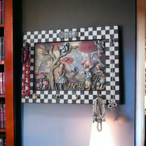 May include: A black and white checkered frame with a whimsical Alice in Wonderland scene. The frame has hooks for hanging jewelry, including a red beaded necklace and a silver chain necklace with a snake charm.