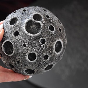 May include: A black and white moon-shaped cake with craters and a white frosting. The cake is being held in a hand.