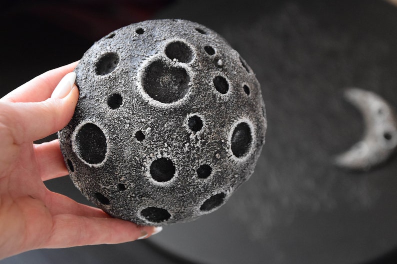 May include: A black and white moon-shaped cake with craters and a white frosting. The cake is being held in a hand.